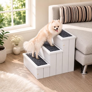 May include: A white three-step pet staircase with dark gray carpeted treads. The structure is made of solid white panels and features small gold-toned hardware accents on the sides, designed to assist small animals in reaching elevated surfaces.