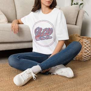 May include: A white short-sleeve t-shirt featuring a circular graphic design with the text 312 and The Chi Love in red and blue. The design includes smaller text arranged in a circle around the central numbers and script lettering.