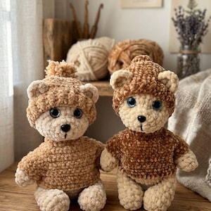 May include: Two handmade crochet teddy bears. The bears are light brown with cream-colored faces, paws, and bellies. One bear wears a hat with a pom-pom, the other wears a sweater. Both have blue eyes and black noses. The bears are sitting on a wooden surface.