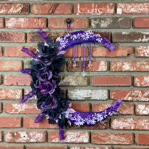Pode incluir: Uma decoração de parede em forma de meia-lua com um fundo de tijolos. A lua é decorada com rosas pretas e roxas, flores brancas e fitas roxas. Fios de contas com pingentes de cristal pendem da lua.