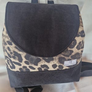 May include: Black backpack with a flap closure and adjustable straps. The flap and base are black, while the front features a tan and black leopard print pattern. A small white label with text is on the side.