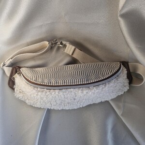 Könnte beinhalten: Eine cremefarbene Bauchtasche mit einem gewebten Oberteil und einem flauschigen weißen Boden. Die Tasche hat einen braunen Reißverschluss und Besatz, mit einem cremefarbenen verstellbaren Riemen und silberfarbener Hardware. Zum Tragen um die Taille oder quer über dem Körper.