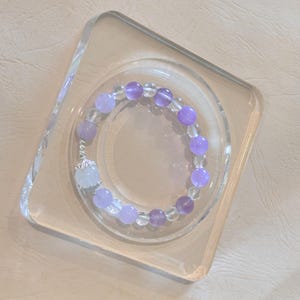 May include: A beaded bracelet featuring alternating clear and purple beads. The bracelet is displayed on a clear, square-shaped stand. The beads vary in shades of purple, creating a gradient effect. A white, ornate charm is also included.
