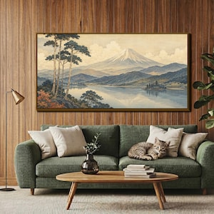 May include: A rectangular wall art print featuring a landscape painting of a snow-capped mountain, a calm lake, and tall pine trees. The artwork is displayed in a thin brown frame and is mounted on a wall with vertical wood paneling above a sofa.