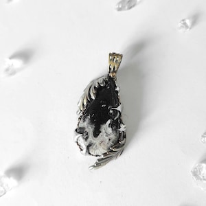 May include: A teardrop-shaped pendant featuring a black and white patterned stone set in a silver-toned metal frame with intricate leaf-like engravings along the edges and a textured bail at the top for attaching to a necklace chain.