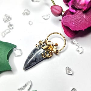 Pode incluir: Um pingente com uma pedra cinzenta polida em forma de lágrima com inclusões escuras, rematado por uma cravação floral em metal dourado. A peça inclui uma argola circular dourada no topo para fixação a uma corrente ou cordão.