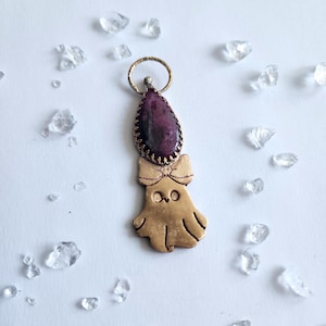 Könnte beinhalten: Handgefertigter Anhänger mit einem goldfarbenen Geisteranhänger mit Schleife, gekrönt von einem tropfenförmigen, dunkelvioletten Edelstein. Der Anhänger hängt an einem kleinen Goldring. Klare Kristalle sind verstreut.