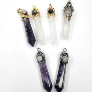Könnte beinhalten: Sechs spitz zulaufende Kristallanhänger in zwei Reihen angeordnet. Die obere Reihe zeigt einen dunkelvioletten Amethyst, zwei klare Quarze mit kleinen runden Akzenten und einen blassrosa Quarz mit silberner Filigrankappe. Die untere Reihe enthält zwei violette Amethystanhänger mit silbernen Filigrankappen.