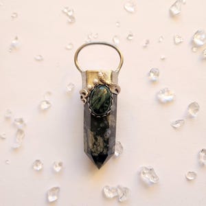 Könnte beinhalten: Silberfarbener Anhänger mit einem dunkelgrünen und schwarzen Kristall. Der Kristall ist in eine silberne Fassung mit einer Schlaufe für eine Kette gefasst. Der Anhänger ist von kleinen, klaren Kristallen auf weißem Hintergrund umgeben.
