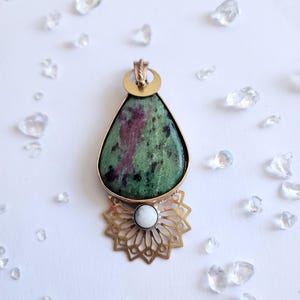 Könnte beinhalten: Ein tropfenförmiger Anhänger mit einem grün-roten Stein in einem goldfarbenen Rahmen. Unter dem Stein befindet sich ein goldfarbenes Filigranmuster mit einem weißen Stein in der Mitte. Klare Kristalle sind auf einer weißen Oberfläche verstreut.