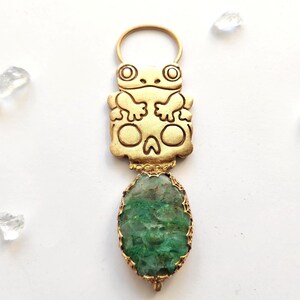 Könnte beinhalten: Ein goldfarbener Metallanhänger mit einer stilisierten Froschfigur an der Oberseite und einer runden Öse. Unter dem Frosch befindet sich ein ovaler, texturierter grüner Stein, der in einen dekorativen goldfarbenen Metallrahmen eingefasst ist.