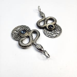 Könnte beinhalten: Zwei Anhänger aus silberfarbenem Metall mit einem gewundenen Schlangendesign und texturierten Schuppen. Jeder Anhänger enthält ein kreisförmiges Filigran-Element und einen dunkelblauen Cabochon-Stein in der Mitte. Oben befindet sich eine Metallöse.