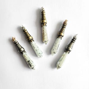 Könnte beinhalten: Fünf Anhänger, die fächerförmig angeordnet sind. Jeder Anhänger hat einen spitz zulaufenden, cremefarbenen, durchscheinenden Körper mit komplizierten schwarzen Blumenmustern, gekrönt von einer strukturierten Metallkappe in Antikmessingoptik.