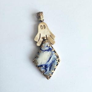Könnte beinhalten: Goldfarbener Anhänger mit einem geisterförmigen Oberteil und einem rautenförmigen Boden. Die Raute hat ein weiß-blaues Marmormuster, das von einer goldenen Filigranarbeit umrahmt wird. Der Anhänger befindet sich auf weißem Hintergrund.