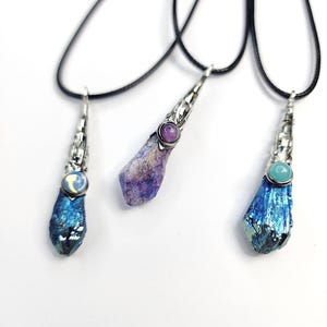 May include: Three pendant necklaces on black cords. Each features a raw, metallic-finish crystal point capped in silver-toned metal. The left pendant has a blue crystal and moon cabochon, the center a purple crystal and purple stone, and the right a blue crystal and teal stone.