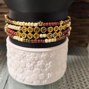 May include: Stack of beaded bracelets with the text "COWGIRLS DON'T CRY" spelled out in gold letter beads. The bracelets are in shades of cream, burgundy, and gold, with small gold crown charms.