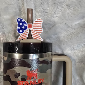 May include: A camouflage-patterned Stanley mug with a red logo and a decorative bow-shaped straw topper. The bow features a patriotic design with red and white stripes and blue stars.