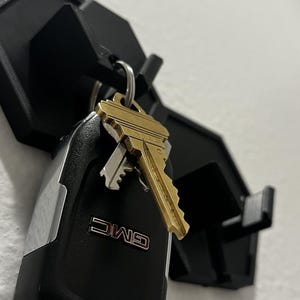 May include: A black key holder mounted on a white wall, holding a set of keys and a GMC key fob. The key holder has a geometric design with multiple hooks. The keys are gold-colored.