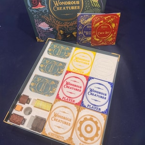Puede incluir: Un juego de mesa Wondrous Creatures que incluye una caja grande ilustrada, dos folletos pequeños y una bandeja de juego con varios mazos de cartas etiquetados como Player, Achievements y Solo, junto a fichas de madera y componentes.