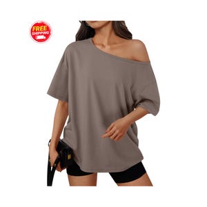 May include: A taupe-colored, off-the-shoulder t-shirt with short sleeves. The shirt has a relaxed fit and is paired with black shorts. A black purse is held in the left hand. The image includes a "Free Shipping" graphic.