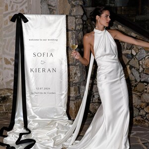 Könnte beinhalten: Ein bodenlanges weißes Satin-Halterkleid mit hohem Ausschnitt und langen, fließenden Stoffbändern. Daneben steht ein hohes Hochzeitsschild aus weißem Stoff mit schwarzem Text: welcome to our wedding, SOFIA & KIERAN, 12.07.2026, La Bastide de Gordes.