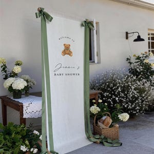 May include: A white baby shower welcome sign with green ribbons and the text "welcome to Jennie's BABY SHOWER" and a teddy bear graphic. The sign is decorated with green ribbons and bows.
