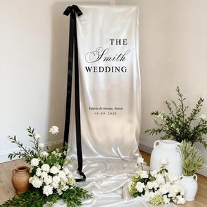 May include: A satin wedding sign with the text "THE Smith WEDDING" and the date 10.09.2025. A black ribbon is tied at the top. White flowers and greenery are arranged around the sign in vases and pots.