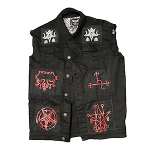 Può includere: Gilet in denim nero con più toppe rosse ricamate con simboli occulti. Il gilet ha una chiusura frontale con bottoni e un colletto. Le toppe includono pentagrammi e altri disegni esoterici. Il gilet è senza maniche.