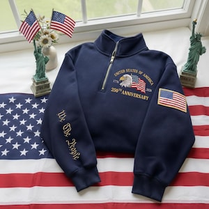May include: Navy blue quarter-zip sweatshirt with "United States of America 250th Anniversary" design, an American flag patch, and "We The People" text. Features a patriotic theme with American flags and Statue of Liberty figurines.