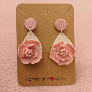 May include: Pink floral earrings with a white teardrop shape and a pink striped circle at the top. The earrings are displayed on a brown card with the text "Handmade with love".