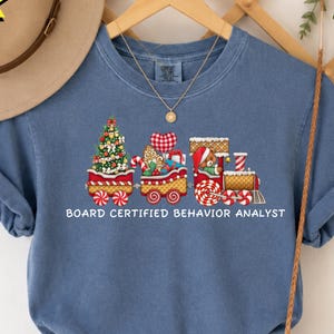 May include: A blue jean colored t-shirt featuring a graphic of a Christmas train carrying a decorated tree, gifts, and a teddy bear. The text BOARD CERTIFIED BEHAVIOR ANALYST is printed in white below the train illustration.