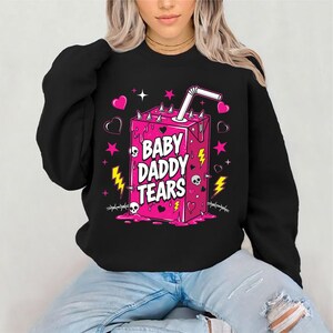 May include: Black sweatshirt featuring a pink juice box graphic with the text "BABY DADDY TEARS." The design includes hearts, stars, lightning bolts, skulls, and barbed wire accents. The juice box has a straw.
