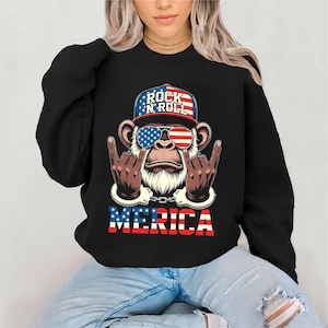 May include: Black sweatshirt featuring a graphic of a monkey wearing sunglasses and a hat with the words "Rock 'N' Roll". The monkey is giving the rock on hand gesture. The word "MERICA" is printed below the monkey.
