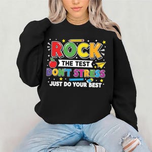 May include: Black sweatshirt with a colorful graphic that reads "ROCK THE TEST DON'T STRESS JUST DO YOUR BEST". The design includes an apple, pencil, paper airplane, and stars. The sweatshirt is worn by a person.