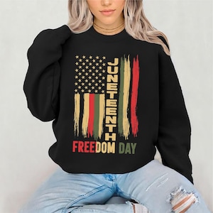 May include: Black sweatshirt featuring a distressed American flag design with the text "Juneteenth Freedom Day" in red, green, and yellow. The flag design includes stars and stripes, celebrating the Juneteenth holiday.