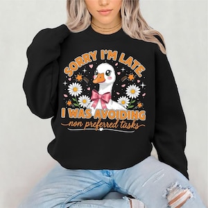 May include: Black sweatshirt with a graphic of a white goose wearing a pink bow, surrounded by daisies and stars. The text reads "SORRY I'M LATE. I WAS AVOIDING non preferred tasks."
