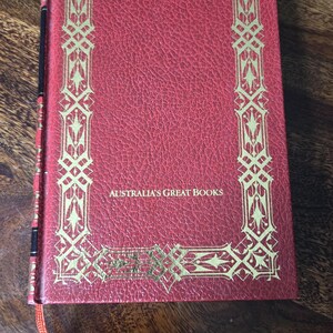 Puede incluir: Un libro rojo con adornos dorados y el texto "AUSTRALIA'S GREAT BOOKS". El libro tiene una cubierta texturizada y una cinta roja como marcador. El libro está sobre una superficie de madera.