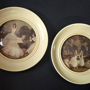 May include: Two cream-colored decorative plates with vintage ballet scenes. The plates have a circular design with a raised edge. The artwork depicts ballerinas in white dresses, set against a backdrop of trees and a garden.