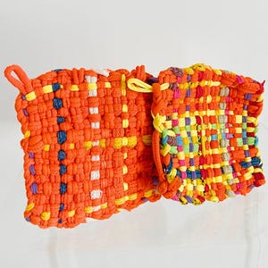 May include: A woven orange pot holder with a loop for hanging. The pot holder features a colorful, textured design with yellow, blue, and white accents. The item is made of woven fabric.