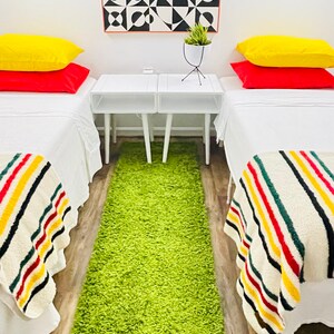 May include: Two twin beds with white bedding and colorful striped blankets. The beds are in a room with white walls and a geometric black and white artwork hanging on the wall. There is a green shag rug on the floor between the beds.