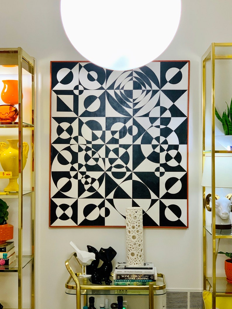 May include: A black and white geometric abstract painting with a red border hangs on a white wall. The painting is surrounded by gold shelves with decorative objects.