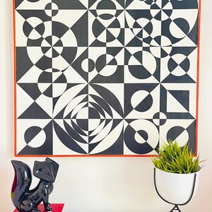 May include: A black and white geometric abstract painting in a red frame hangs on a white wall above a white table with a black fox figurine and a yellow tray.