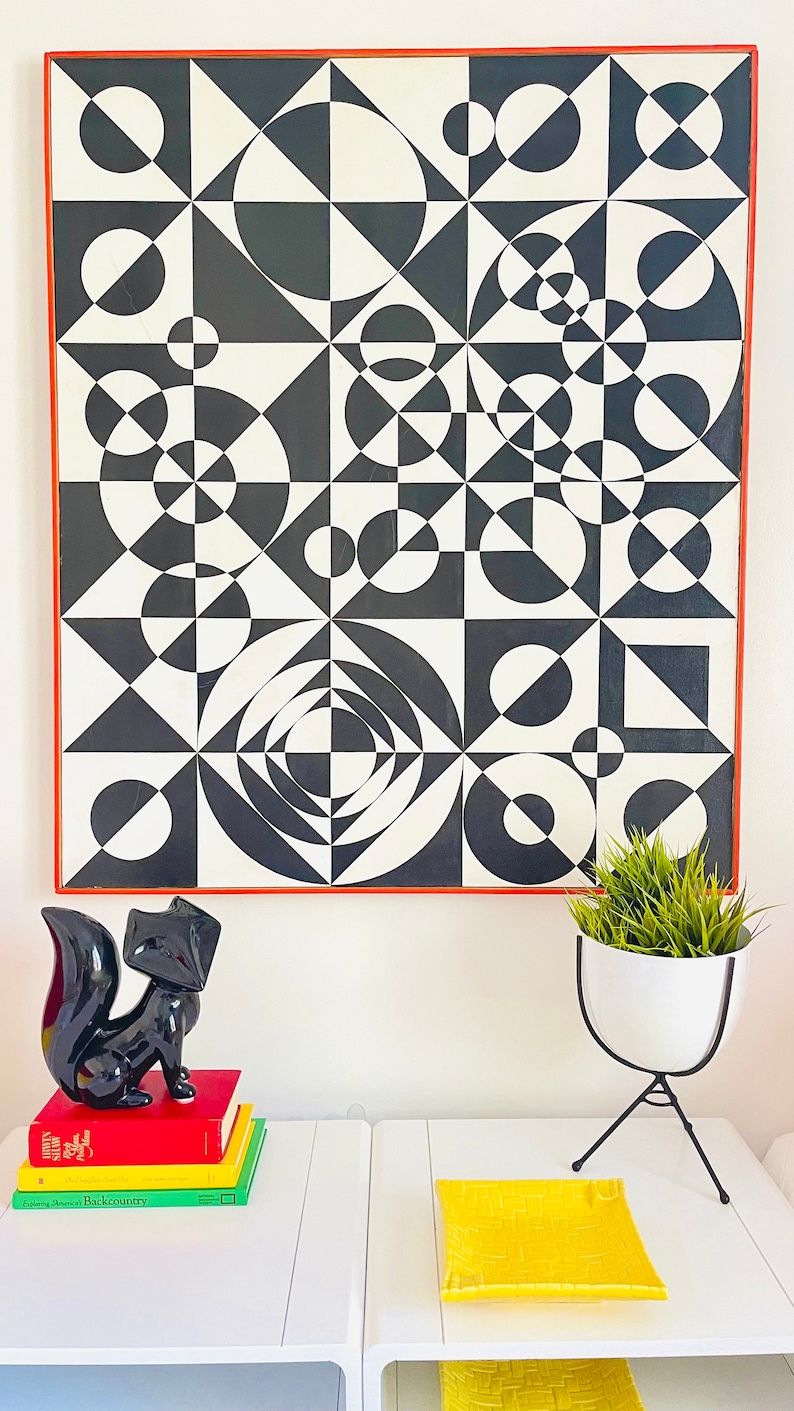 May include: A black and white geometric abstract painting in a red frame hangs on a white wall above a white table with a black fox figurine and a yellow tray.
