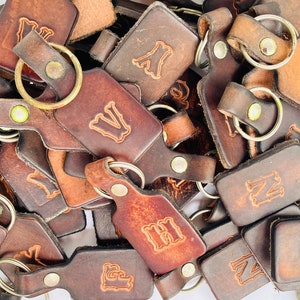 May include: A pile of brown leather keychains with metal rings. Each keychain has a different letter embossed on it.