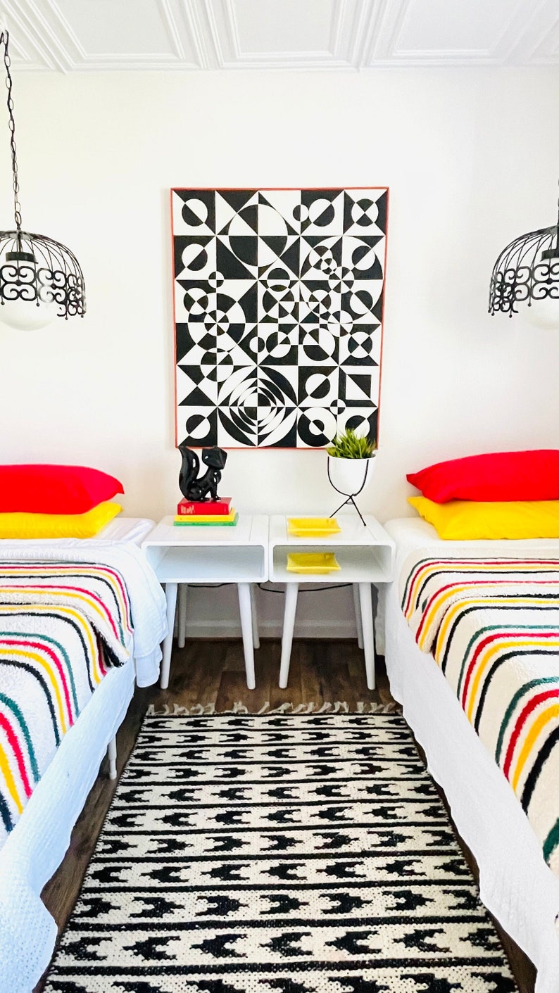 May include: Two twin beds with white linens and colorful striped blankets. The beds have red and yellow pillows. The beds are in a room with white walls and a black and white geometric art print on the wall. There are two white nightstands with black legs and a black squirrel figurine on one of the nightstands.