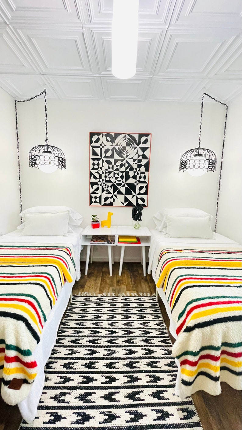 May include: Two twin beds with white bedding and colorful striped blankets. The beds are in a room with white walls and a white ceiling with square tiles. There is a black and white geometric artwork on the wall above a white table with two lamps hanging from the ceiling.