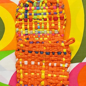 May include: Two woven pot holders in orange, yellow, blue, and pink. The pot holders are made of woven fabric and have a loop for hanging. The background is a colorful, retro-style pattern.