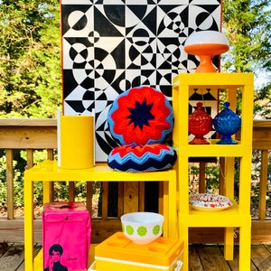 May include: Yellow mid-century modern style end tables with a black and white geometric print artwork, a crocheted pillow, a pink cooler bag, a white bowl with green flowers, and a yellow cooler with the text "SunPacker".