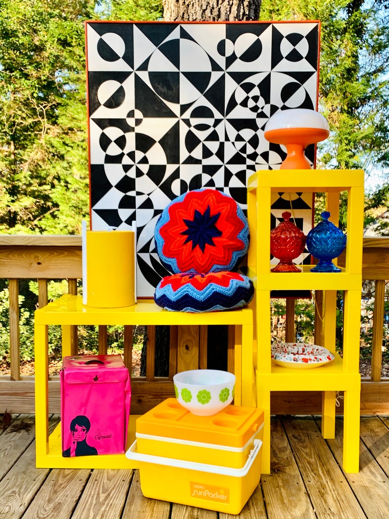 May include: Yellow mid-century modern style end tables with a black and white geometric print artwork, a crocheted pillow, a pink cooler bag, a white bowl with green flowers, and a yellow cooler with the text "SunPacker".