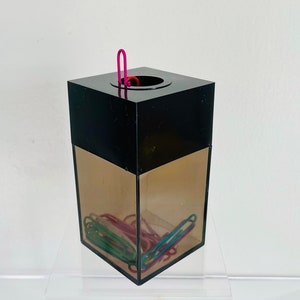 May include: A black plastic container with a clear front panel filled with colorful paper clips. A pink paper clip is sticking out of the top opening.